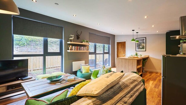 A modern living room with a sofa, a coffee table, and a TV. There are large windows, a bookshelf, and a kitchen area visible. - Host Stay Forest Green Lodge (Alnwick)