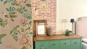 Une pièce avec un mur en briques, une commode verte aux poignées en laiton, un lustre et un tableau encadré posé sur la commode.
