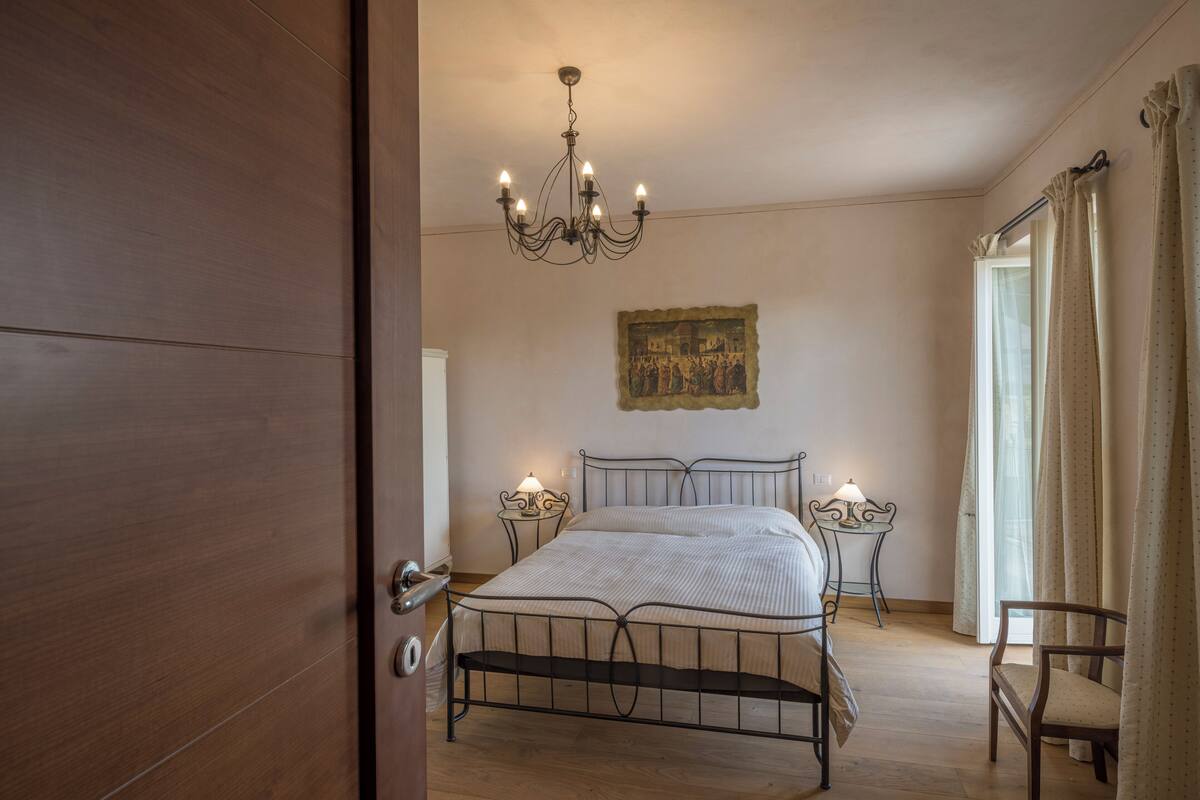 Une chambre à coucher avec un lit en métal, un lustre, une table de chevet avec une lampe et une fenêtre avec des rideaux.