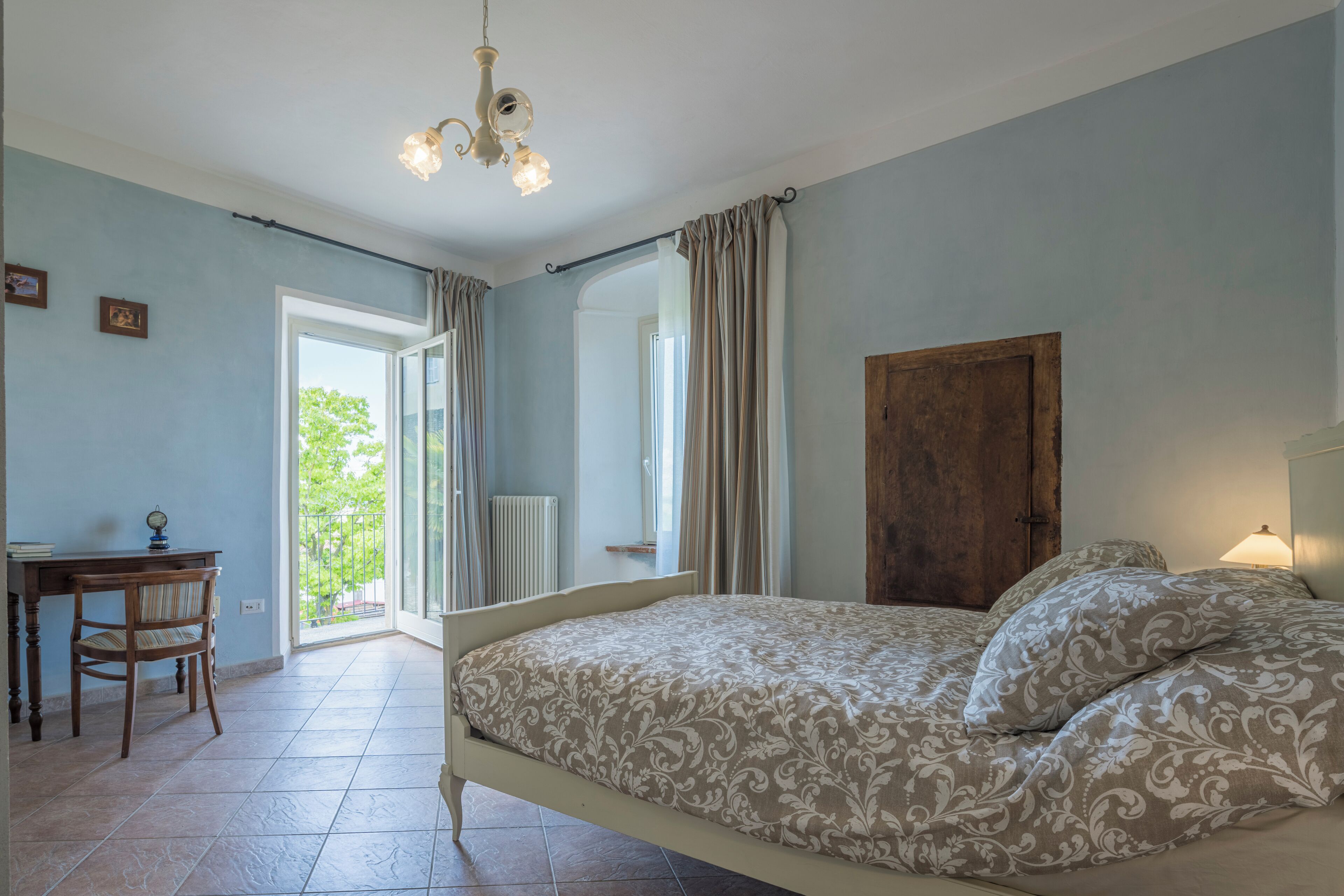 A bedroom with a bed, a desk, and a chair. There is a window with curtains and a view of greenery outside.