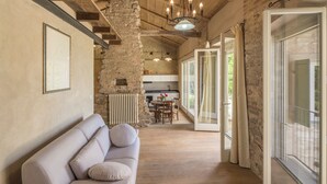 A spacious living area with a beige sofa, wooden flooring, stone walls, and a view of the kitchen and outdoor space. - Arpusè Relax nelle Langhe Barolo with Pool (Monforte d'Alba)