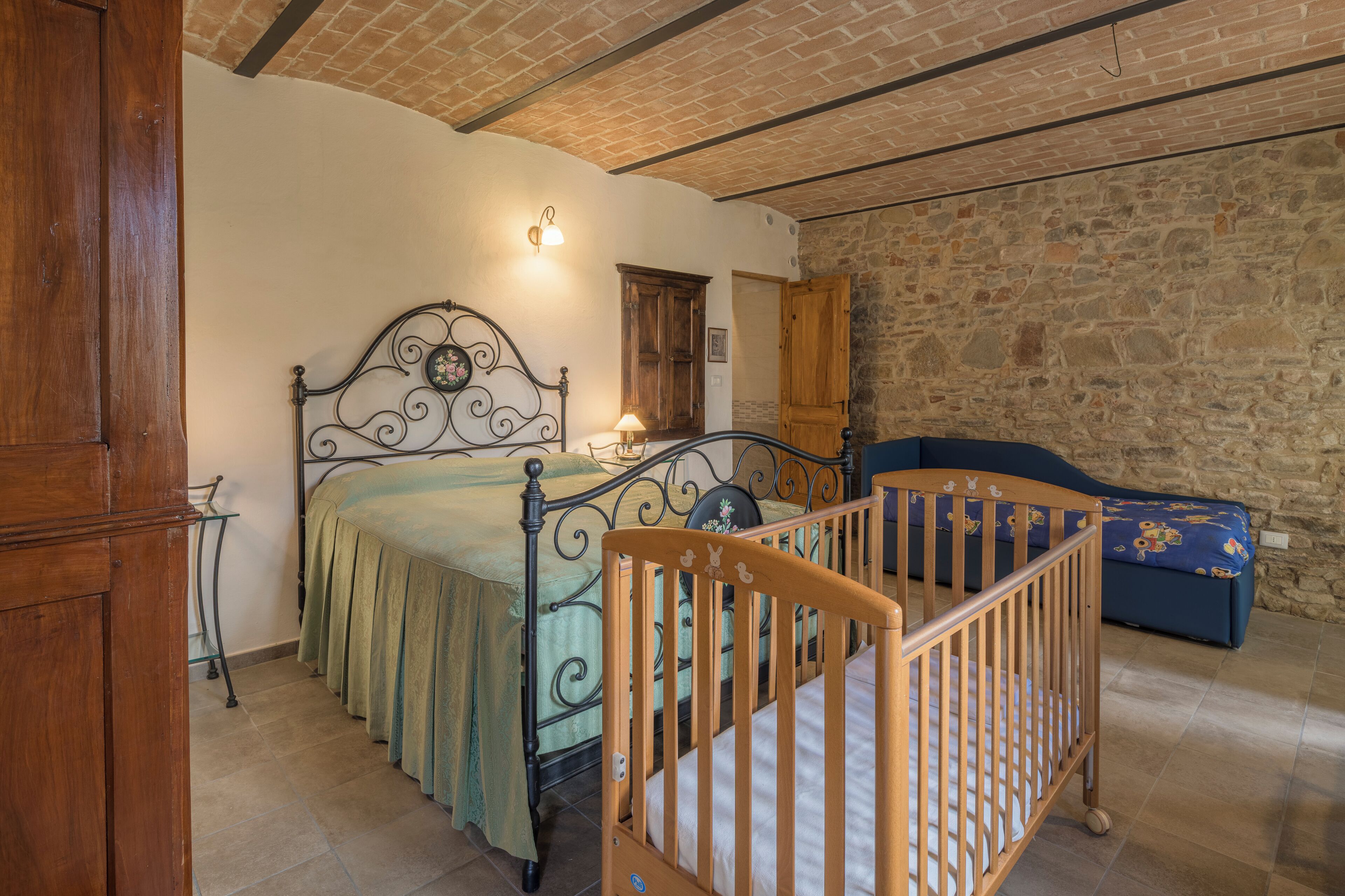 A bedroom with a wooden crib, a metal bed frame, a stone wall, and a wooden door.