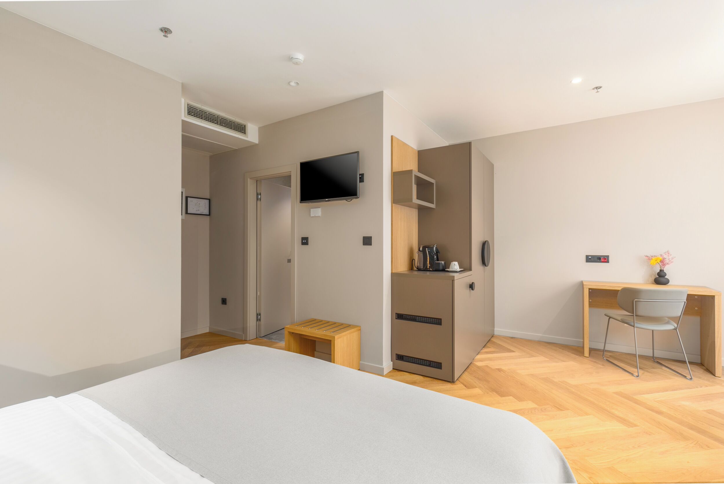 A modern hotel room with a large bed, a desk with a chair, a television, and a small kitchenette.