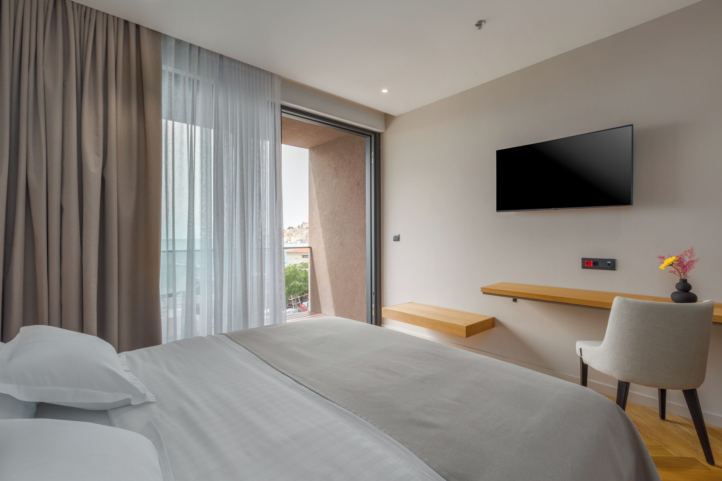 Una habitación de hotel moderna con una cama grande, un televisor montado en la pared, un escritorio con una silla y una ventana con cortinas.