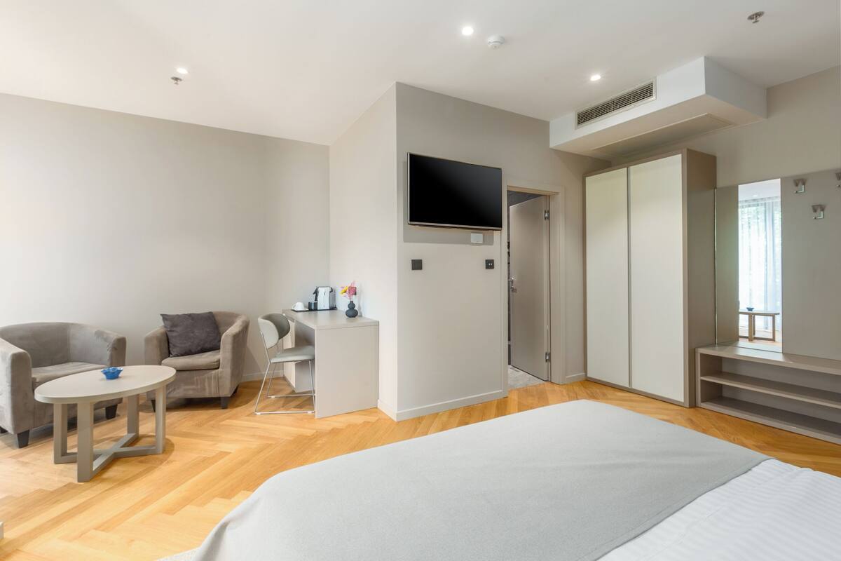 A modern hotel room with a bed, a desk, two armchairs, a TV, and a wardrobe.