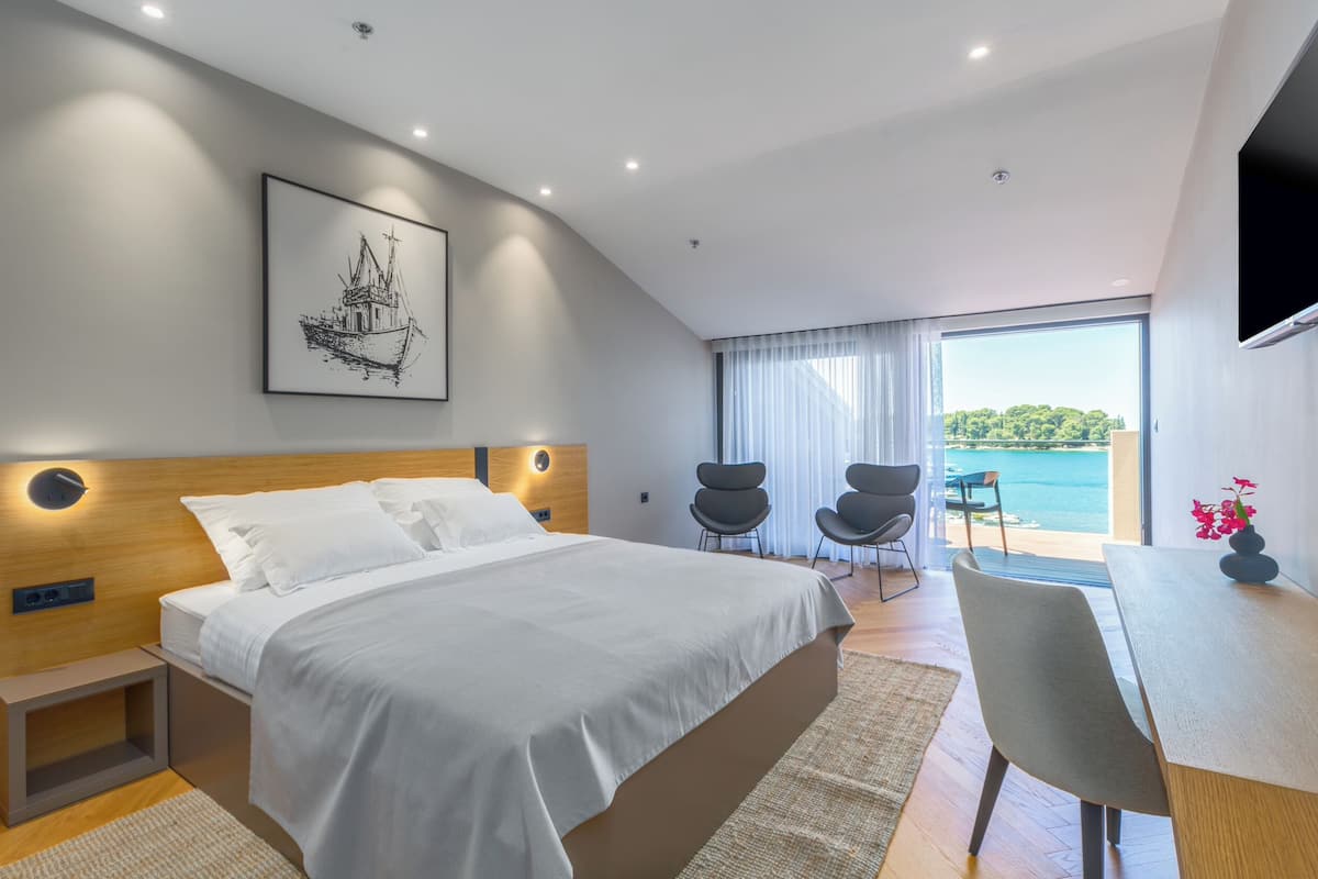 A modern bedroom with a large bed, a desk, and a chair, a large window with a view of the sea, and a framed picture on the wall.