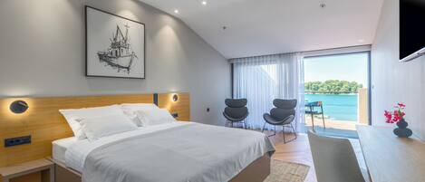 A modern bedroom with a large bed, a desk, and a chair, a large window with a view of the sea, and a framed picture on the wall.