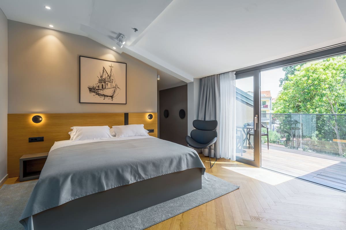 A modern bedroom with a large bed, a wooden headboard, a framed picture, a chair, and a balcony with a view of greenery.