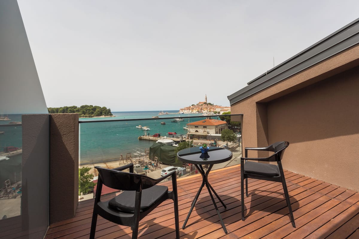 A balcony with two chairs and a small table, overlooking a coastal town with boats and a historic building.