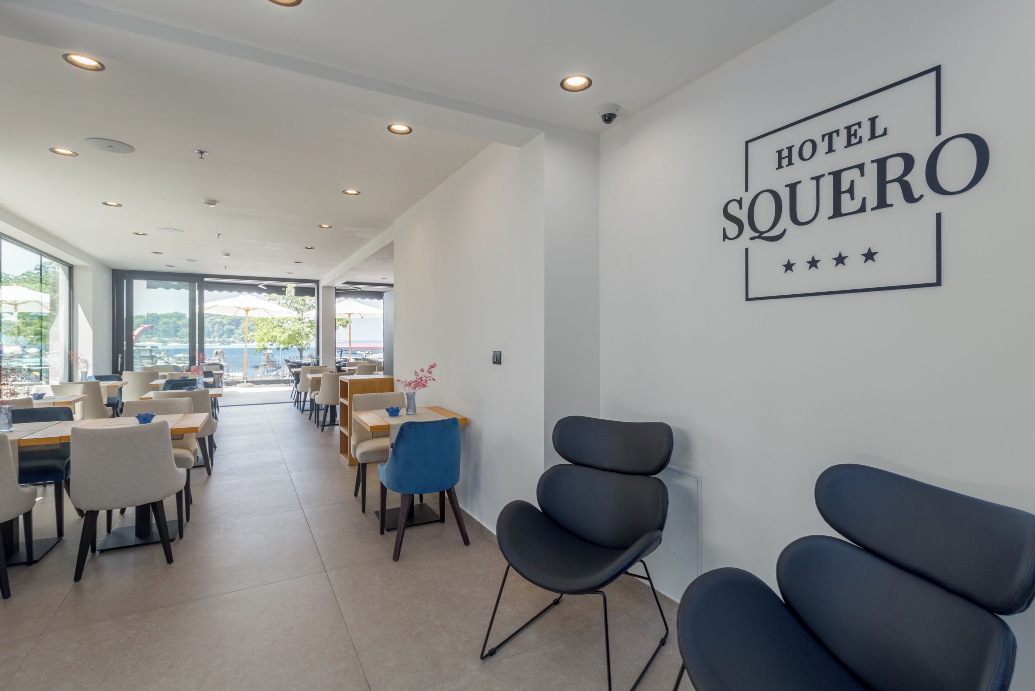 A modern hotel lobby with a large window, a wall sign reading "HOTEL SQUERO," and several seating areas with tables and chairs.