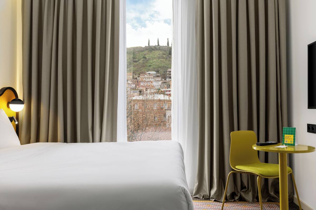 A hotel room with a bed, a chair, a small table, and a view of a cityscape through large curtains.