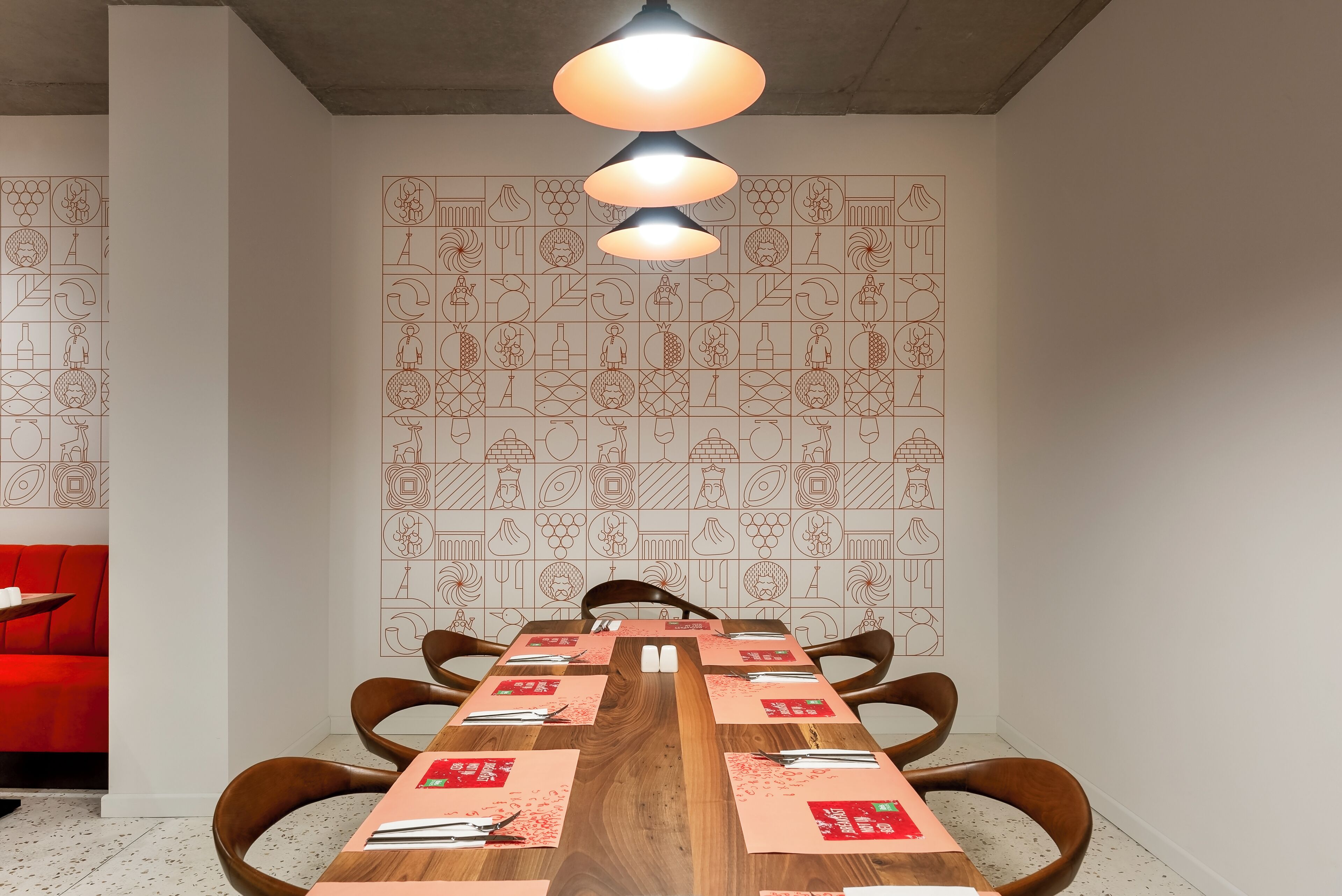 Un espace repas avec une longue table en bois, des chaises à l’assise rembourrée de couleur rouge et un mur orné d’un motif géométrique.