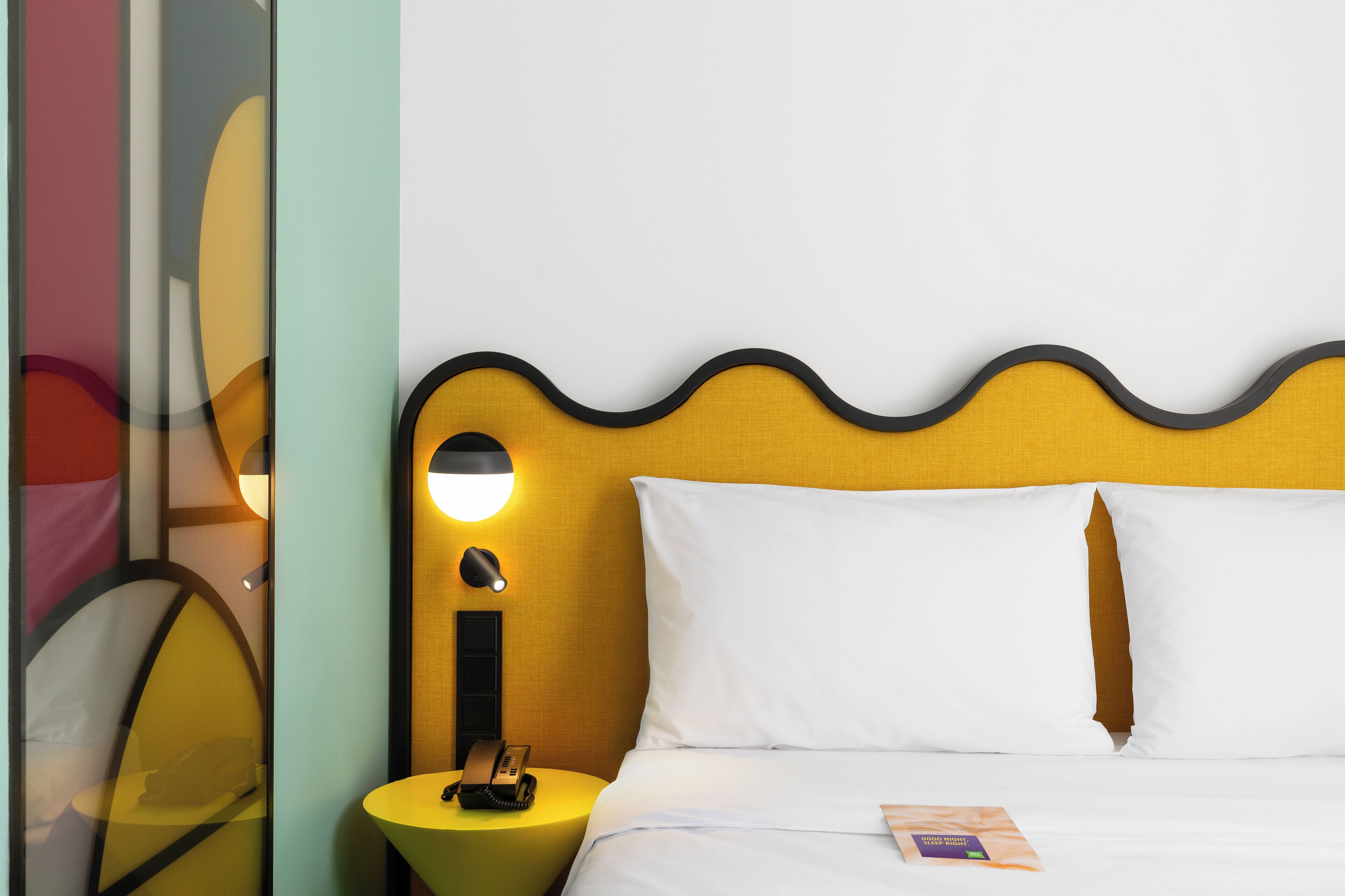 A neatly made bed with white linens, a single pillow, and a small bedside table with a lamp and a telephone.
