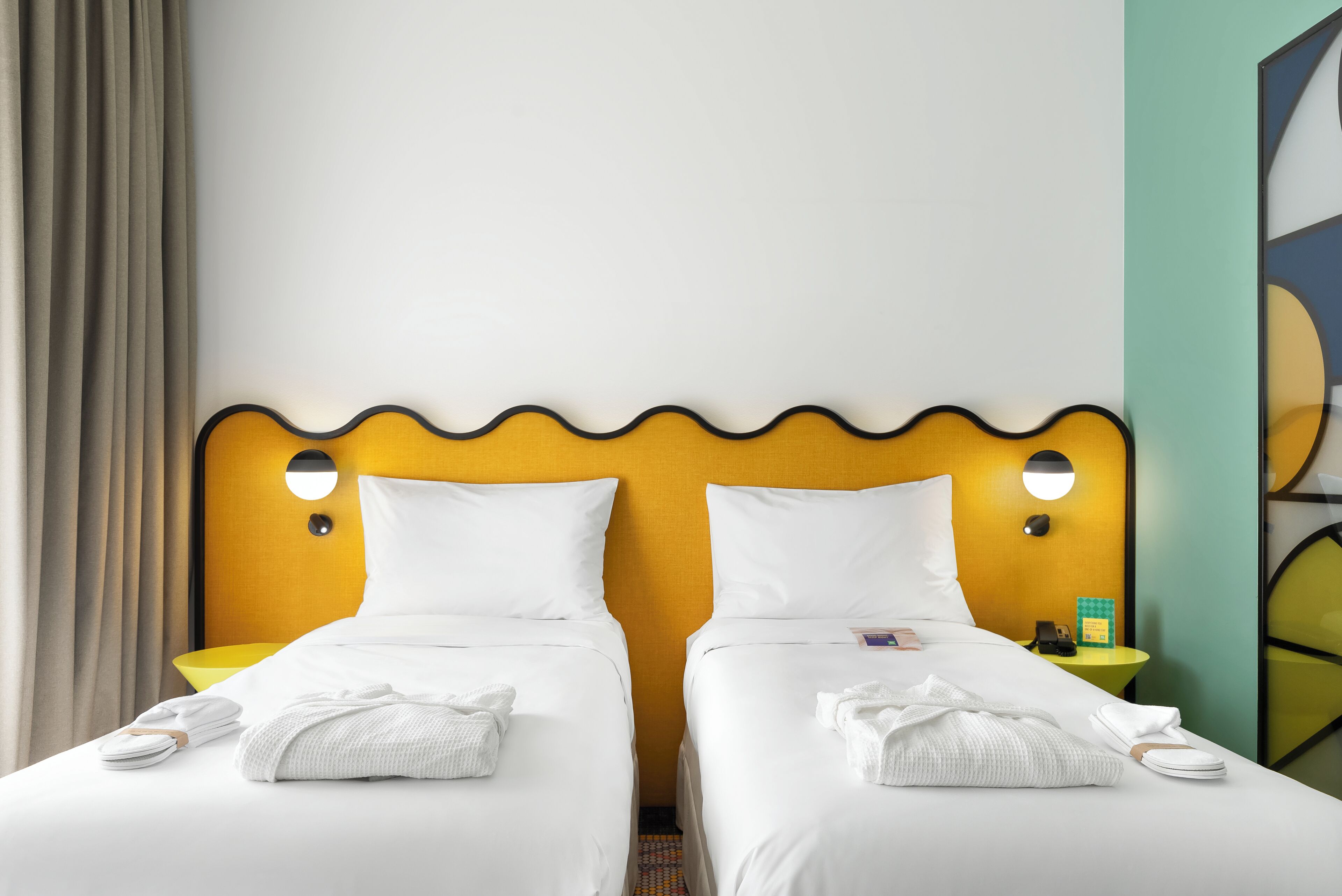 Ein Hotelzimmer mit zwei Betten, jeweils mit weißen Laken und Handtüchern, einem gelben Kopfteil und einem kleinen Nachttisch mit Lampe.