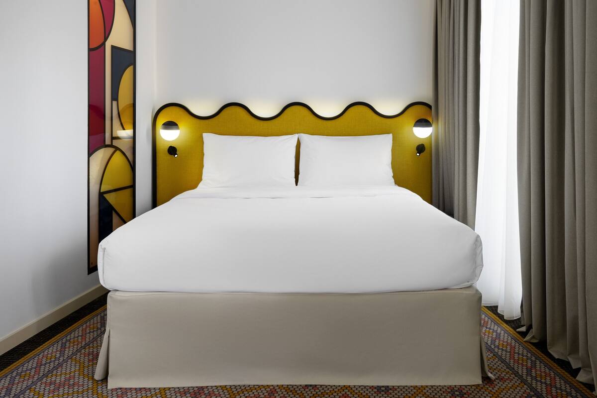 A hotel room with a large bed, a headboard with integrated lighting, a decorative wall piece, and a patterned carpet.