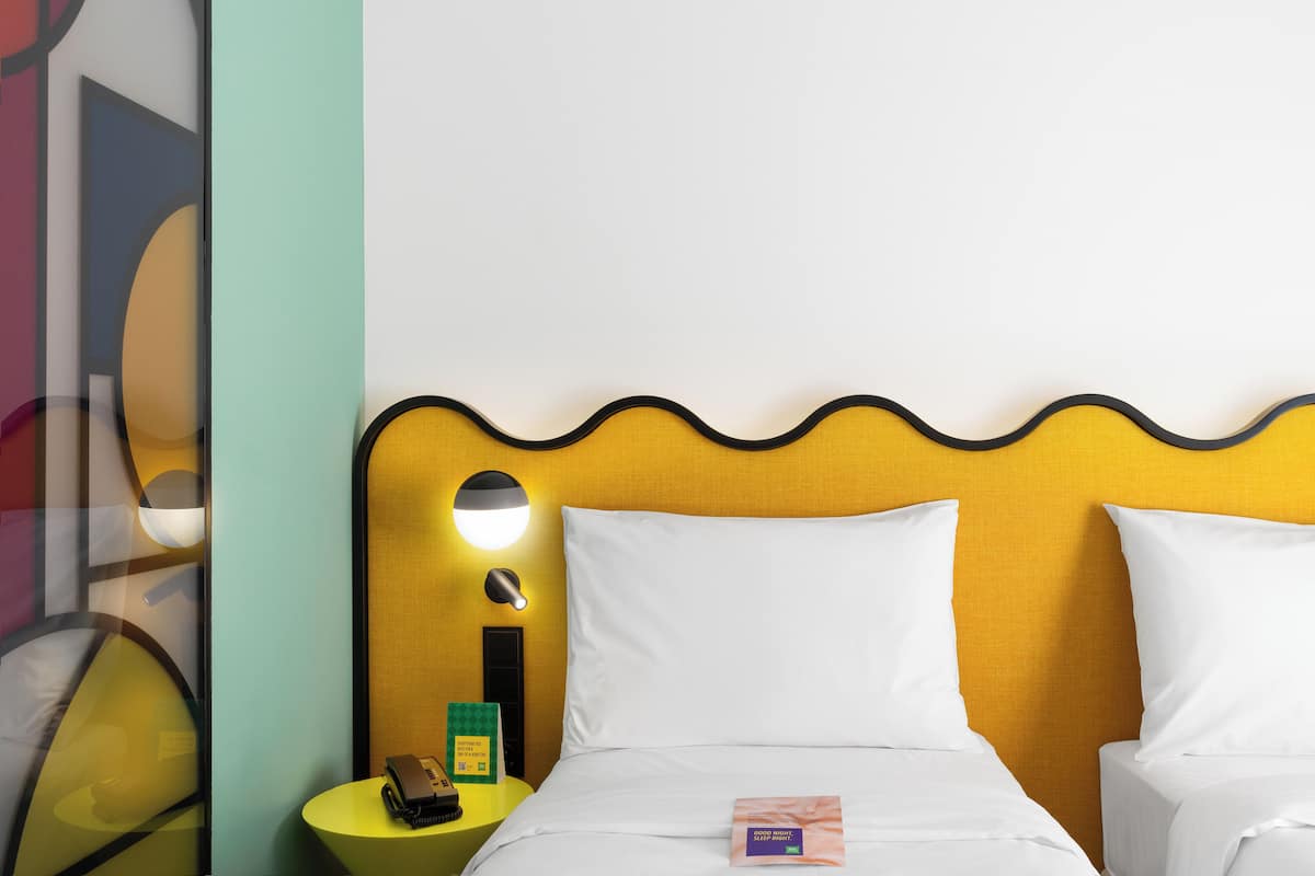 A hotel room with a yellow headboard, white bedding, a bedside table with a lamp, and a nightstand with a phone and a green card.