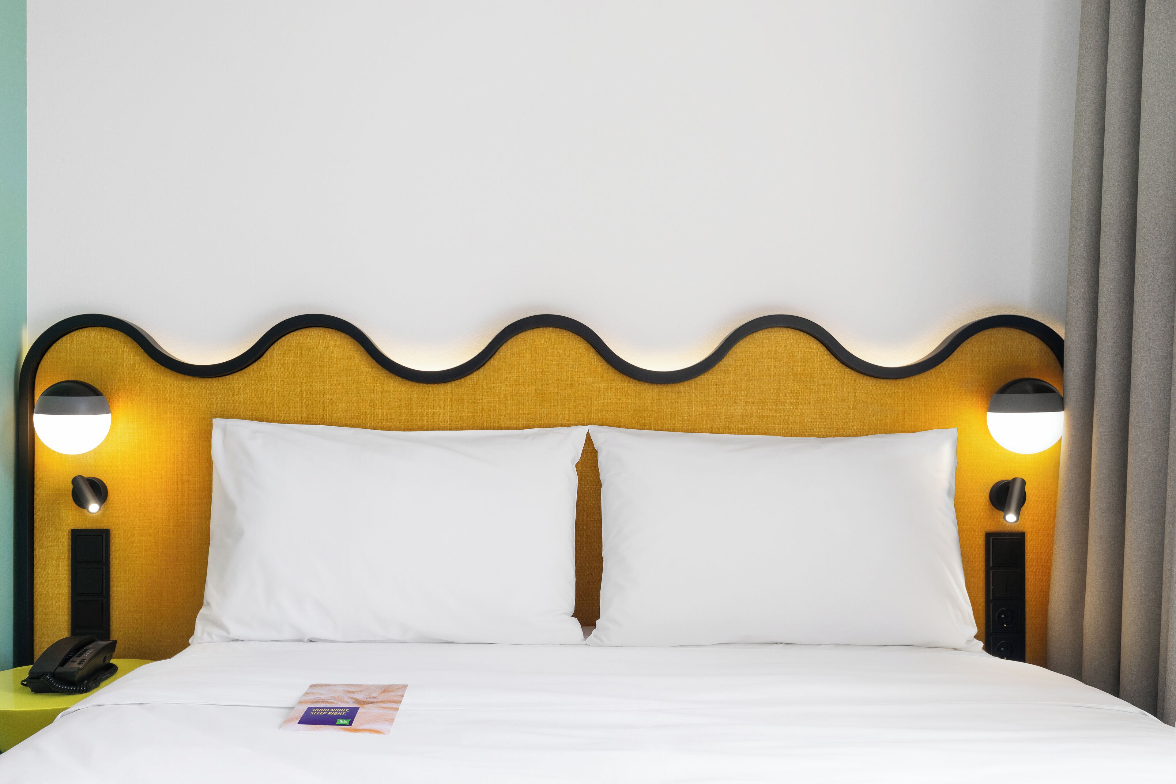 A hotel room with a bed, white bedding, a yellow headboard, and two wall-mounted lamps.