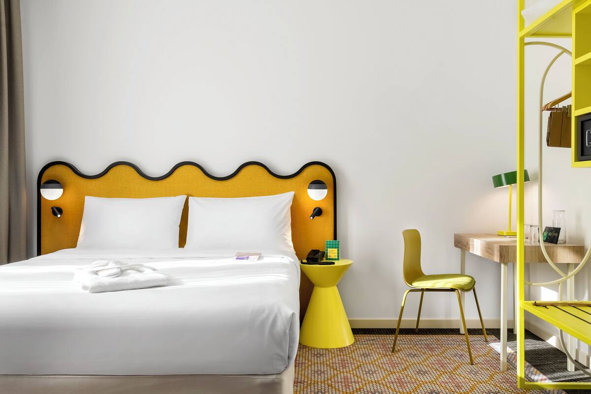 A hotel room with a bed, a yellow side table, a yellow chair, a wooden nightstand, and a yellow shelving unit.