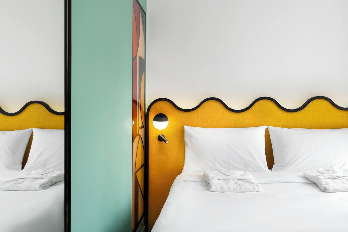 A neatly made bed with white linens and two folded white towels, a yellow headboard with a wavy design, and a wall-mounted light fixture.