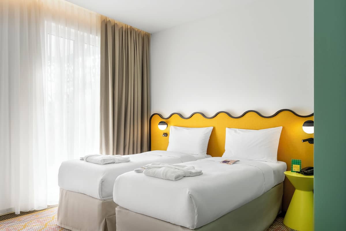 A hotel room with two beds, a yellow accent wall, a small bedside table with a phone and a green object, and a window with sheer curtains.