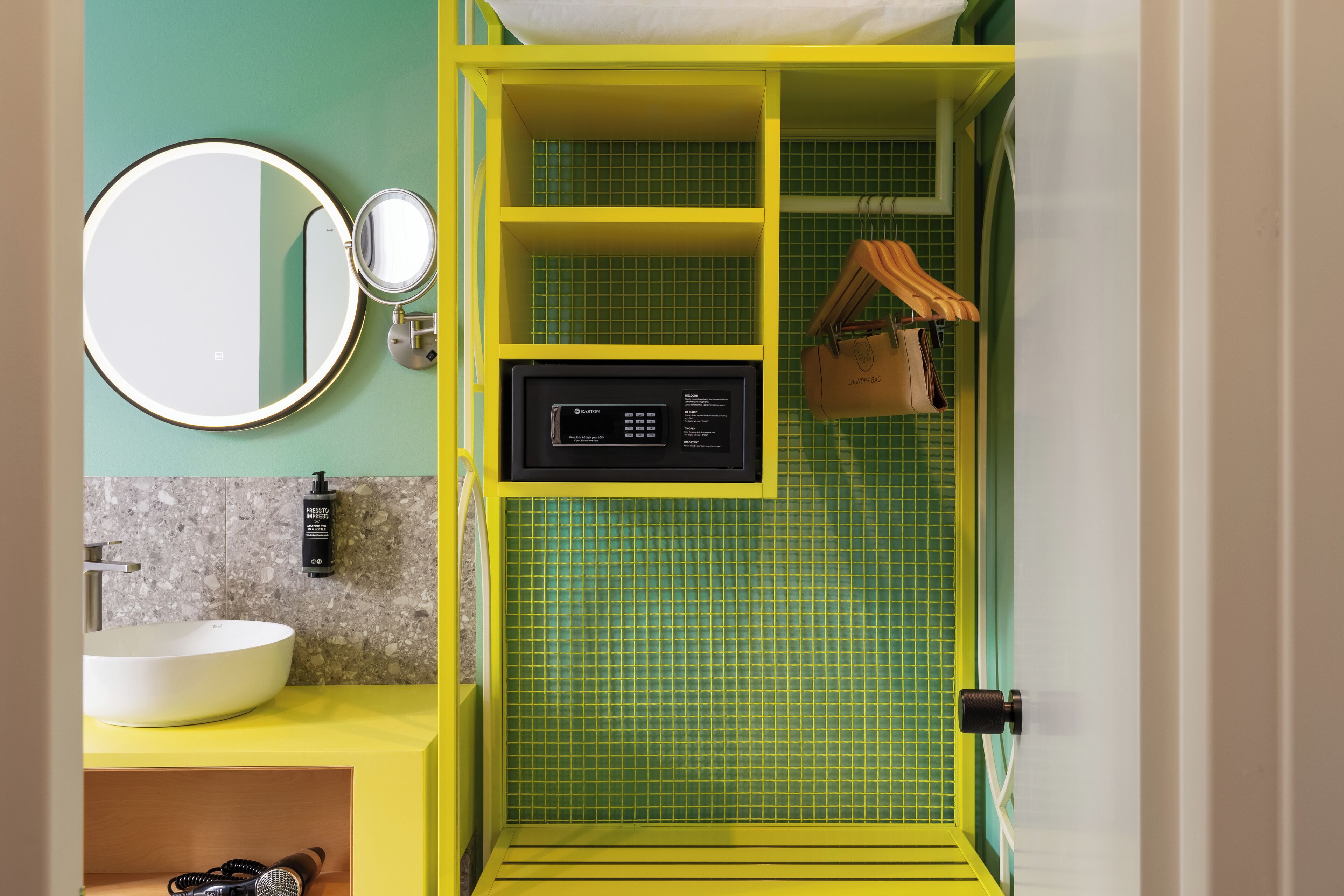 A bathroom with a yellow vanity, a round mirror, a built-in heater, and a green tiled shower area.