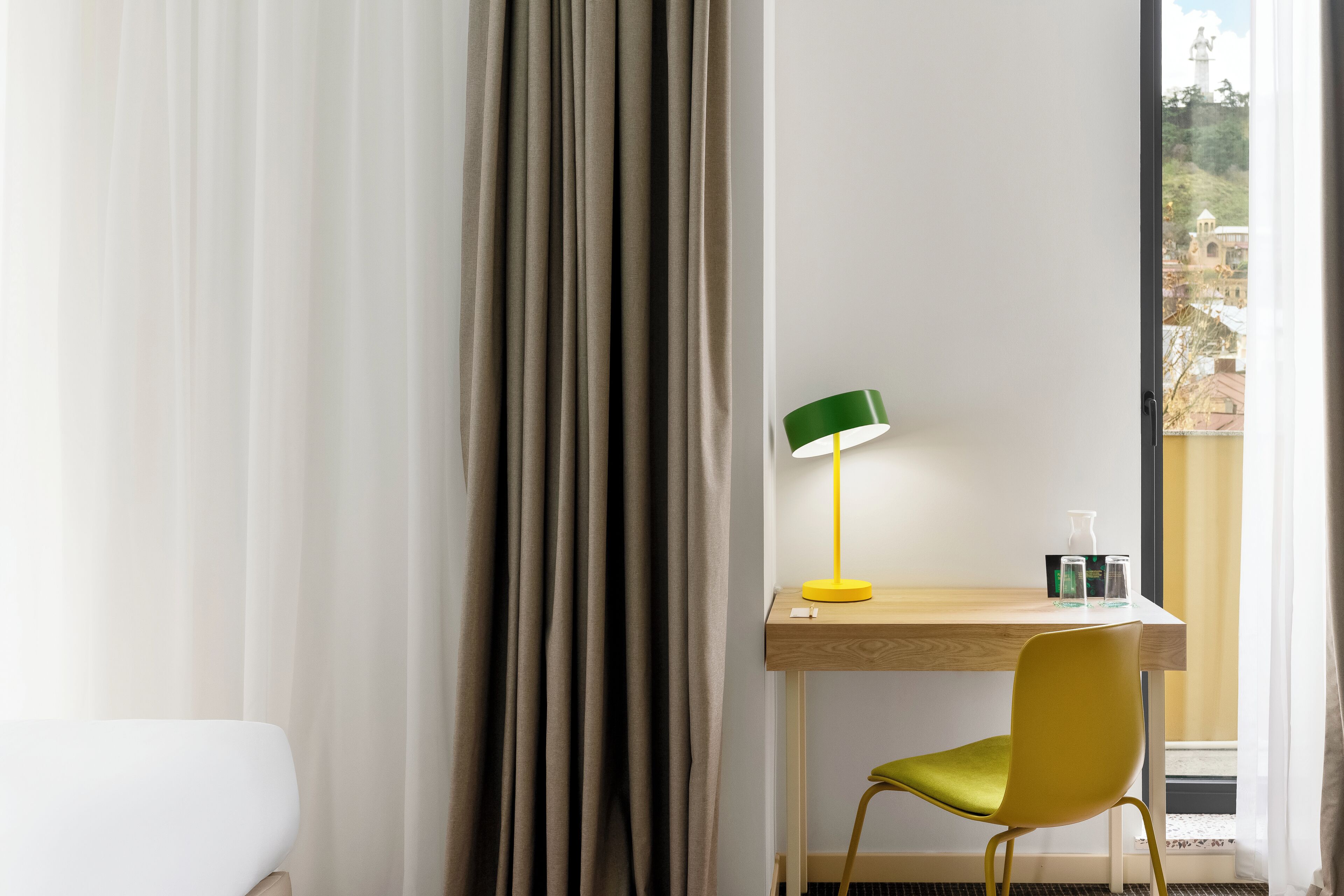 A hotel room with a bed, a desk with a lamp, a chair, and a view of a building through the window.