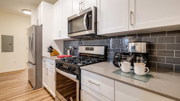 A modern kitchen with white cabinets, a stainless steel refrigerator, a microwave, a stove, and a coffee maker. - Gorgeous Apt with Kitchen (Lexington)