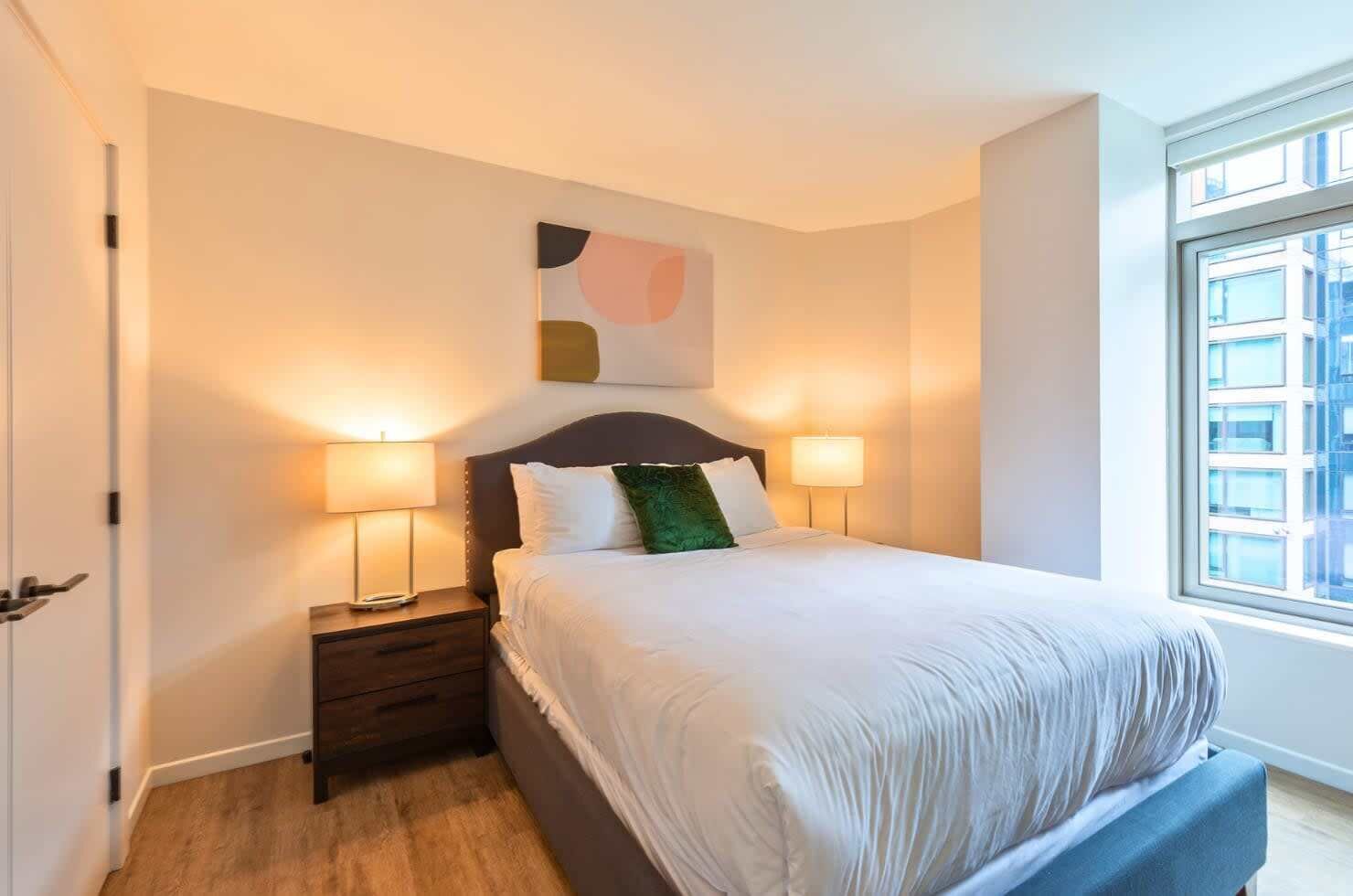 A neatly arranged bedroom with a large bed, two bedside lamps, a nightstand, and a window with a view of buildings.