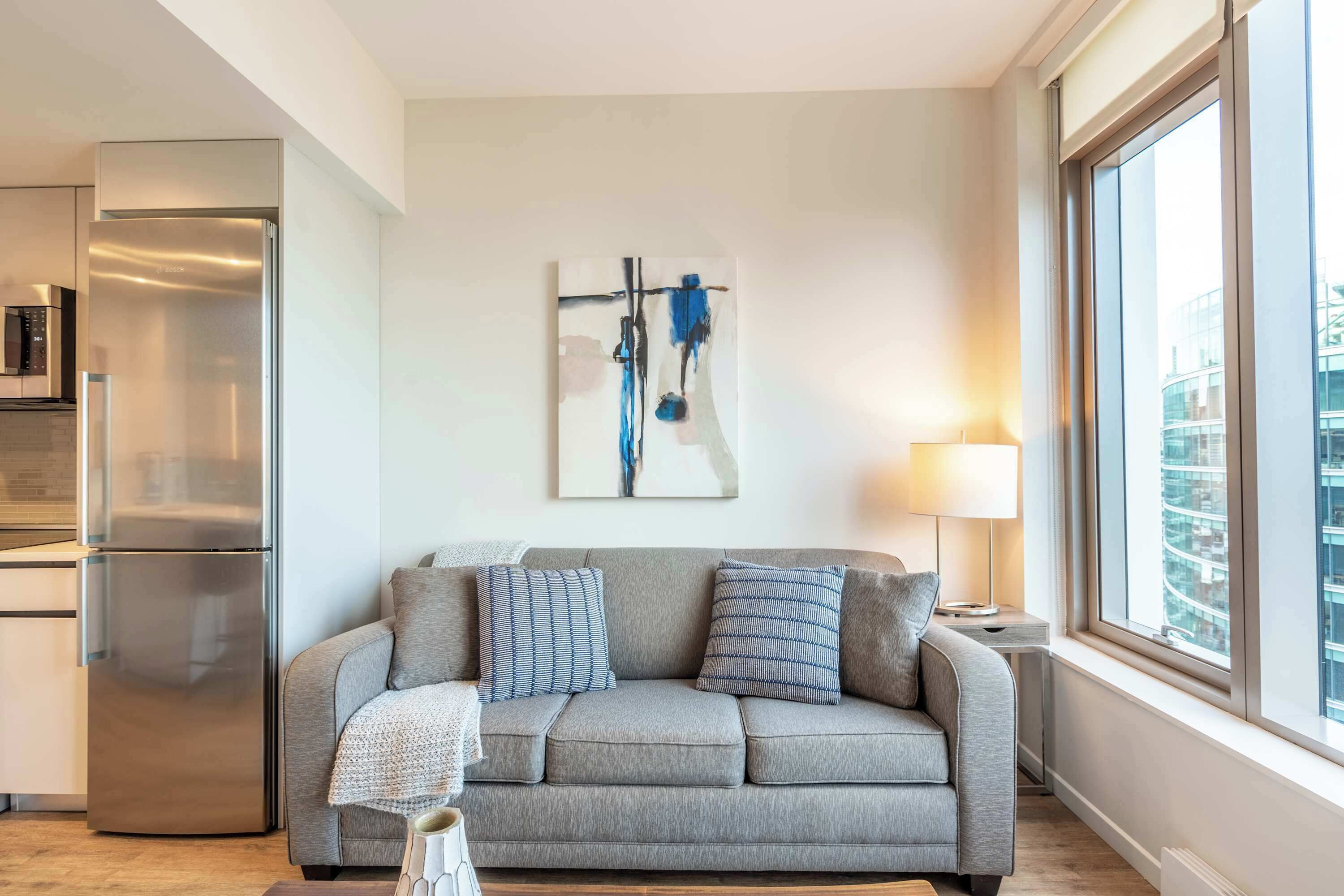 Una sala de estar moderna con un sofá gris, un refrigerador de acero inoxidable y un gran ventanal con vistas a la ciudad.