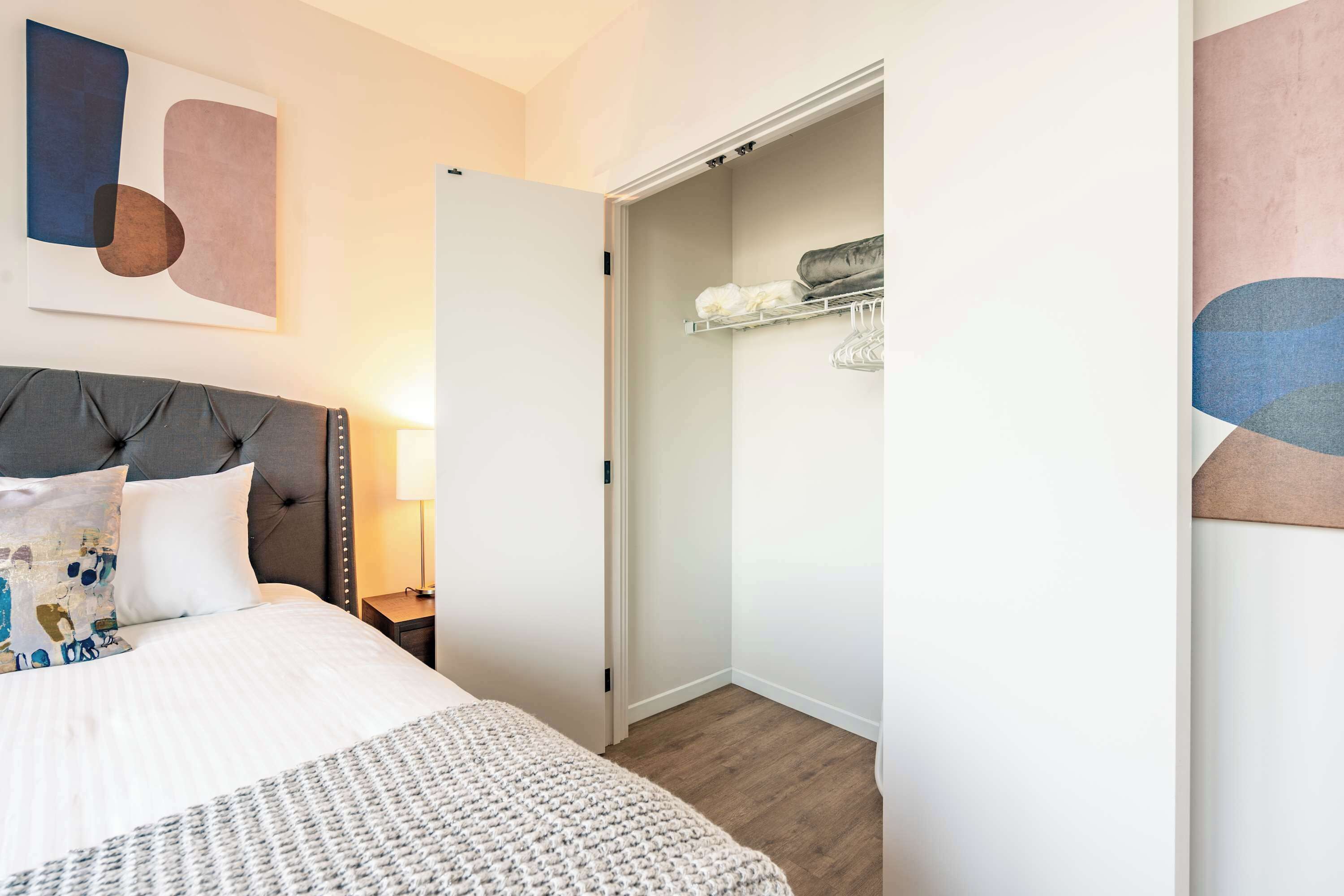 A neatly arranged bedroom with a tufted headboard, a large bed, a bedside table with a lamp, and a closet with shelves.