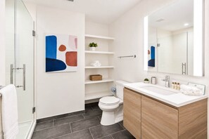 Une salle de bain moderne avec une douche à parois de verre, des toilettes blanches, un meuble-lavabo en bois et une étagère murale où sont rangés les articles de toilette.