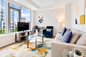 Un salon moderne avec une grande fenêtre, un téléviseur à écran plat, une table basse en verre, un fauteuil bleu, un canapé gris et un lampadaire.