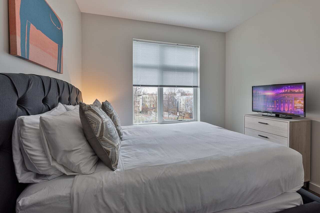 A bedroom with a large bed, a television on a wooden side table, and a window with blinds.