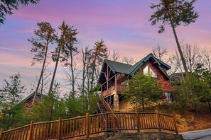 A cabin with a wooden deck and a staircase leading to a second-story balcony, surrounded by trees. - A Cozy Retreat: Unwind, Relax, Repeat (Sevierville)