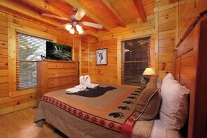 A wooden cabin bedroom with a bed, a ceiling fan, a television, and a window with blinds. - A Cozy Retreat: Unwind, Relax, Repeat (Sevierville)