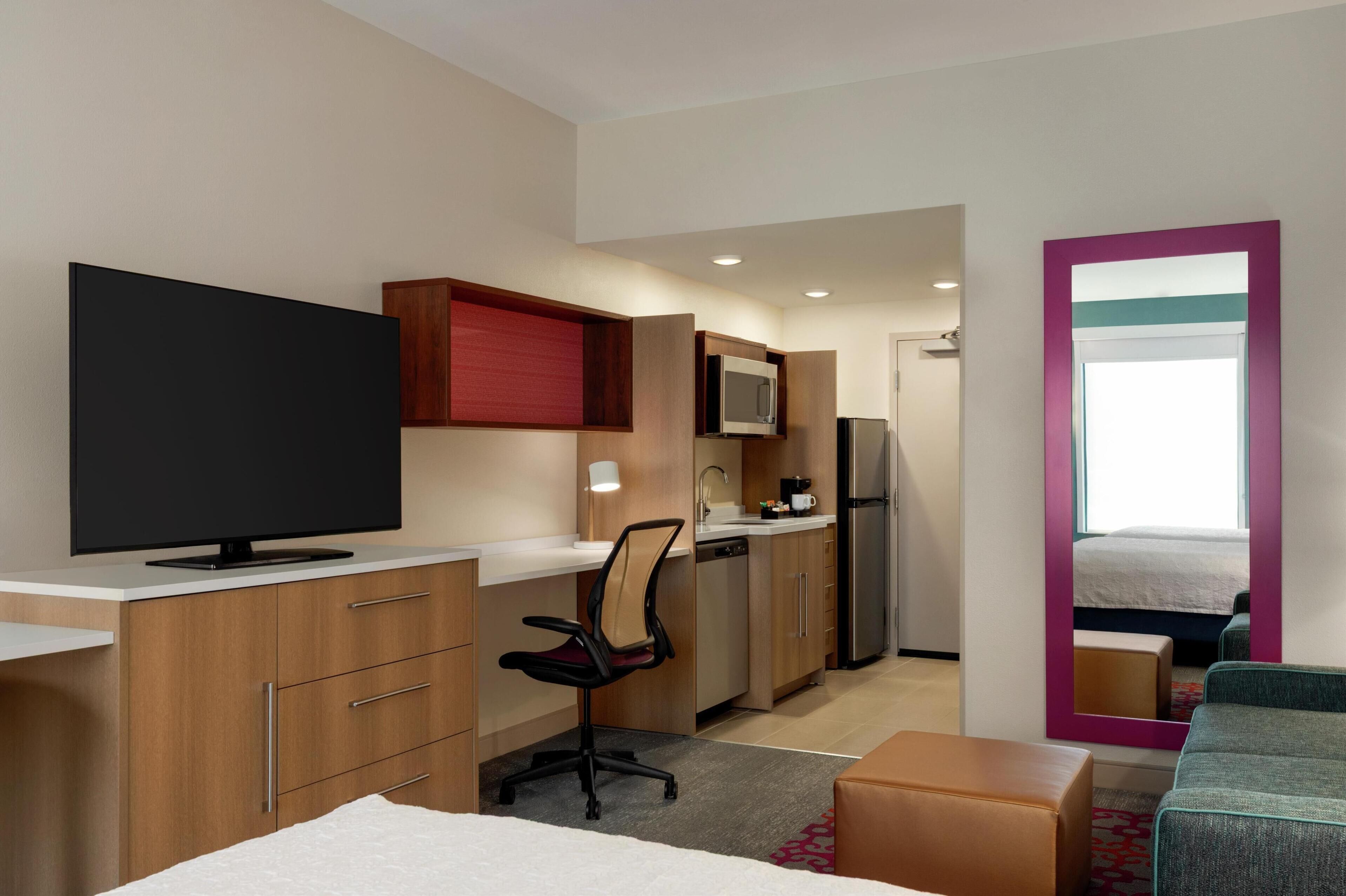 A compact hotel room with a kitchenette, a bed, a TV, a desk with a chair, and a large mirror.