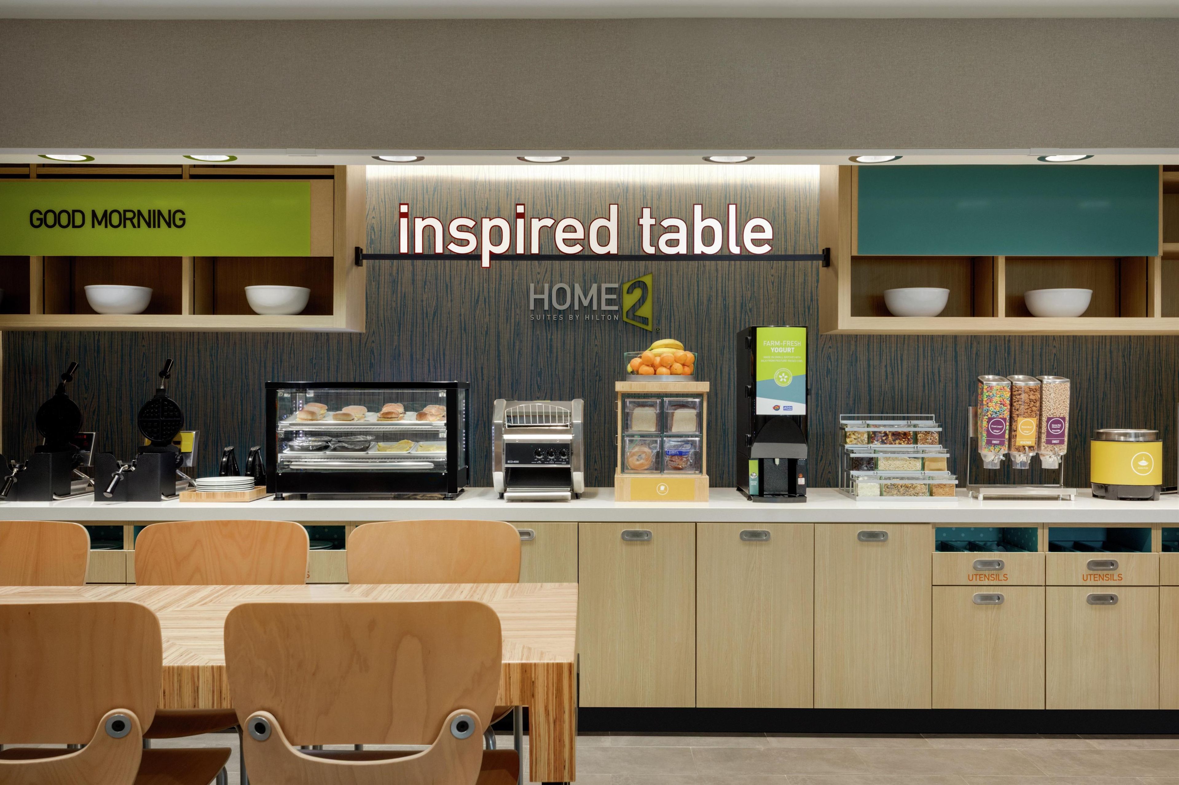 Un área de cocina moderna con una barra desayunadora, identificada como “mesa inspirada”, y un cartel que dice “BUENOS DÍAS”.