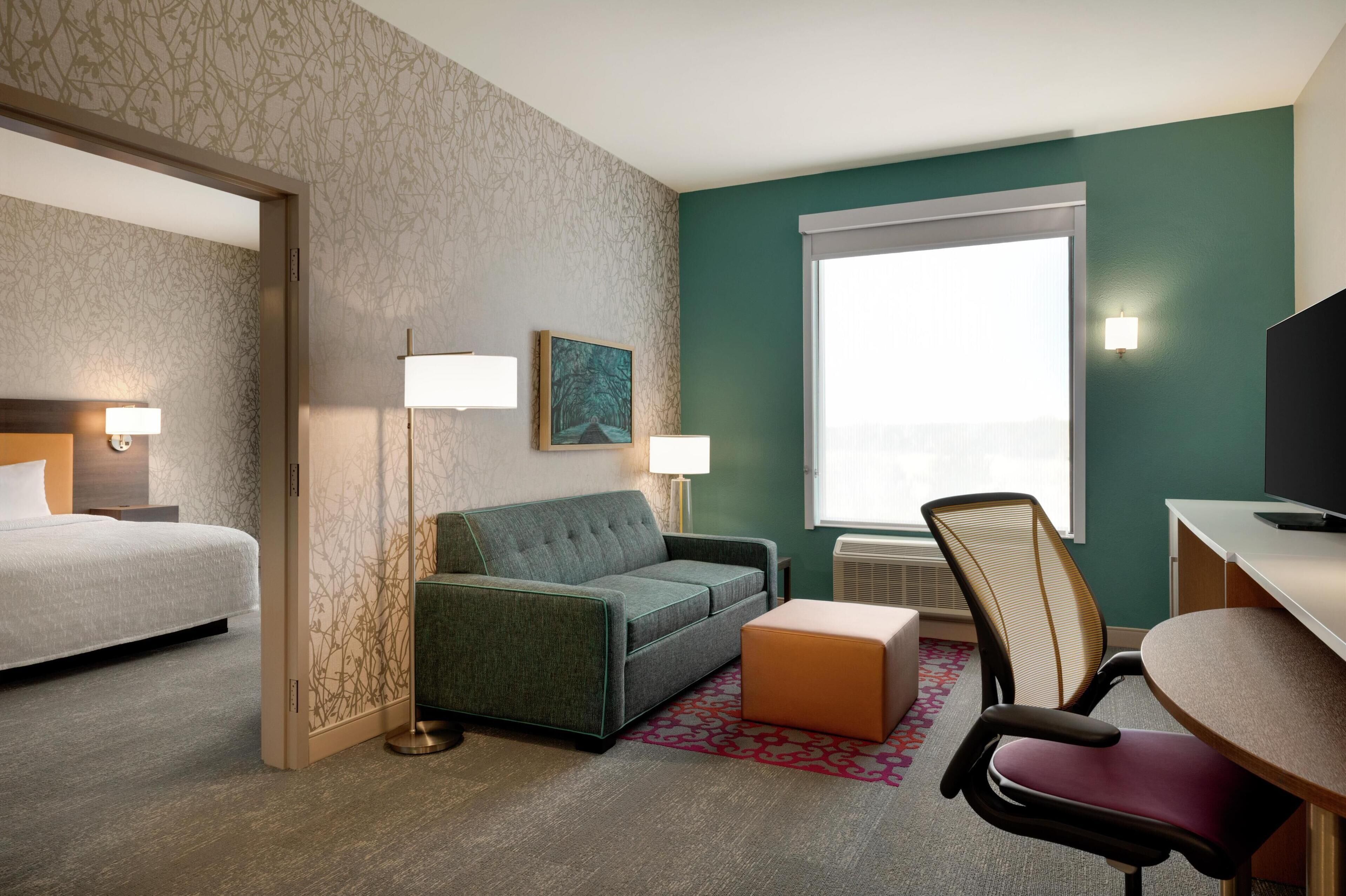 A hotel room with a bed, a sofa, a desk, and a chair. There is a window with a view, a painting on the wall, and a lamp.