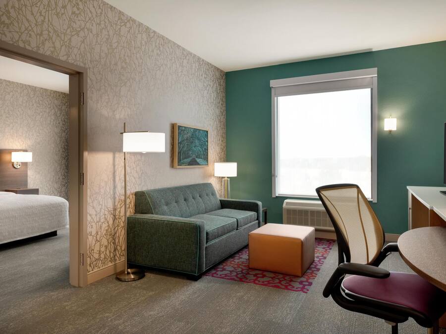 A hotel room with a bed, a sofa, a desk, and a chair. There is a window with a view, a painting on the wall, and a lamp.
