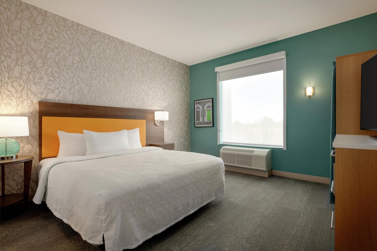 A hotel room with a bed, a nightstand, a lamp, a window with blinds, and a wall-mounted TV.