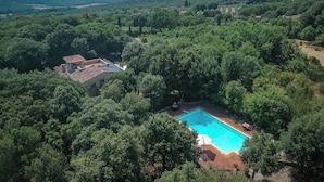 An aerial view of a property with a swimming pool surrounded by dense trees, a small building with a tiled roof, and distant hills. - Santa Colomba House Nature And Pool (Monteriggioni)