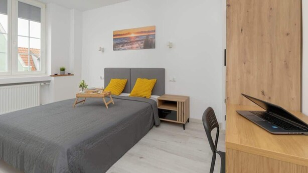 Ein Schlafzimmer mit einem Bett, einem Schreibtisch mit Laptop, einem Nachttisch und einem Bild an der Wand.