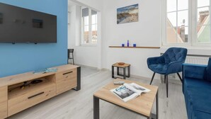 Ein modernes Wohnzimmer mit blauer Wand, einem hölzernen TV-Schrank, einem blauen Sofa, einem kleinen Tisch mit einer Zeitschrift und einem Fenster mit Blick auf Gebäude.