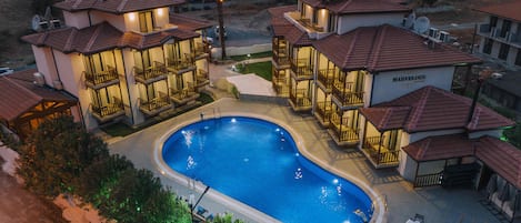 Um resort com piscina central rodeada por edifícios de vários andares com varandas, um letreiro indicando 'MARVELOUS HOTEL' e uma antena parabólica em um dos edifícios.