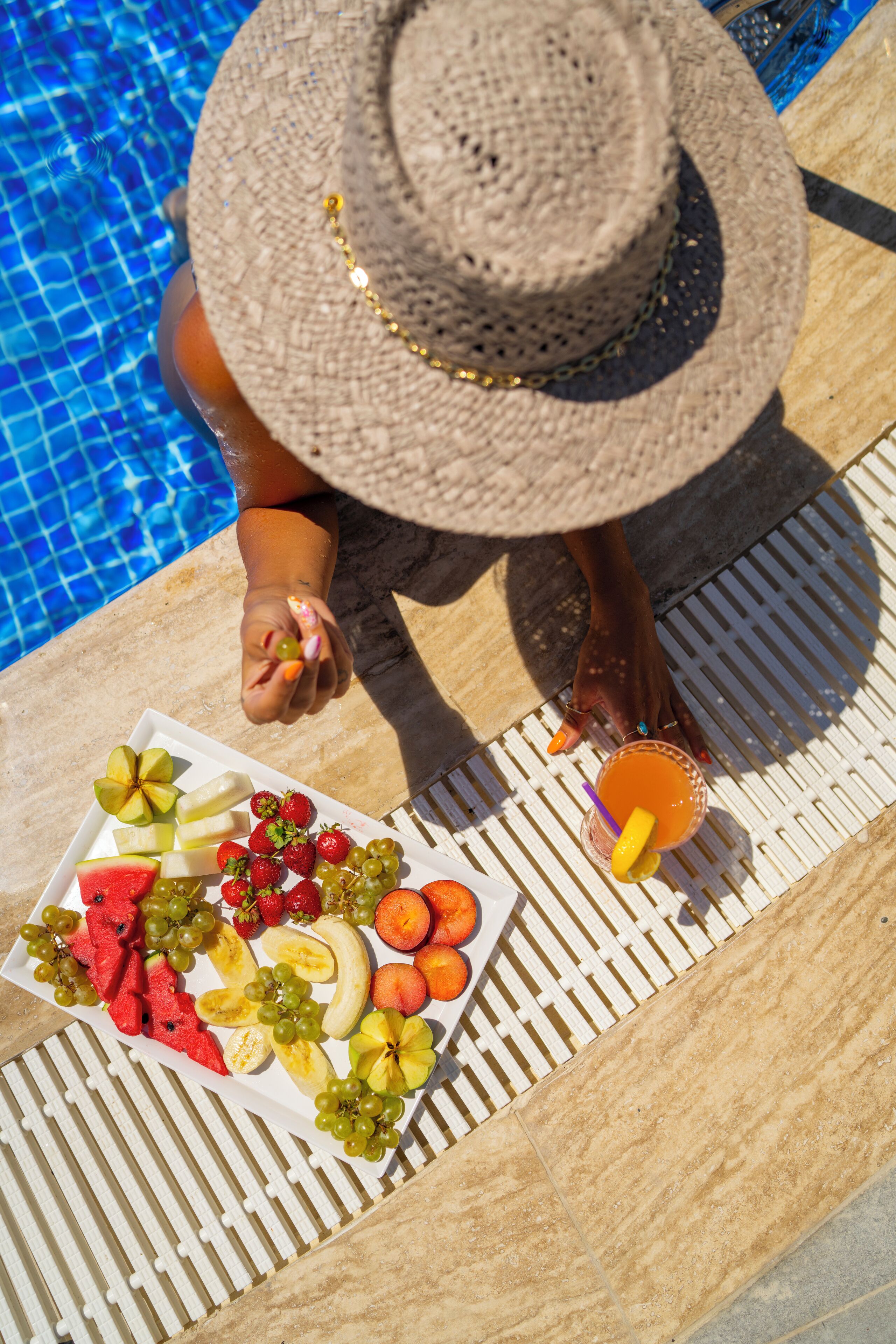 A person relaxing by a poolside with a plate of fruit and a drink.