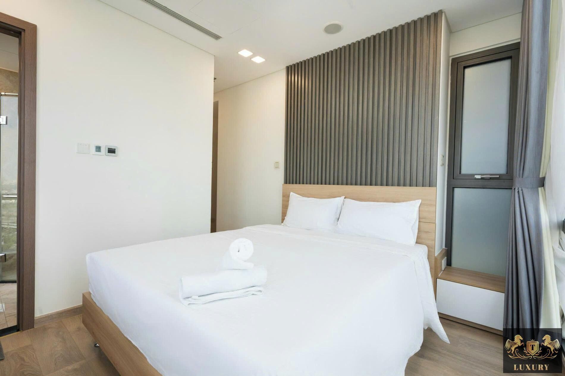A hotel room with a large bed, a wooden headboard, a window with curtains, and a view of the city.