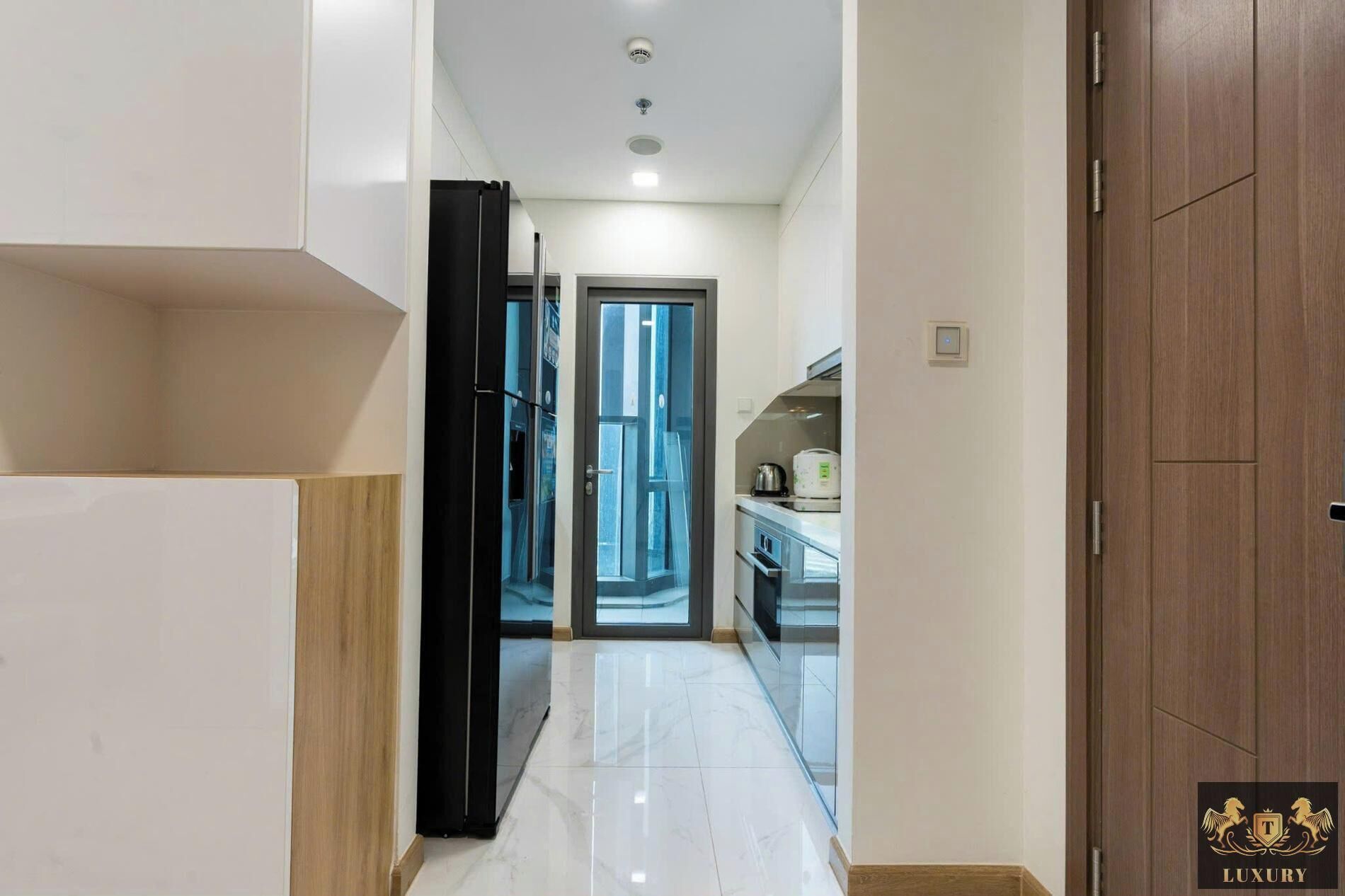 A hallway leading to a kitchen with a modern design, featuring a black refrigerator, a glass door, and a wooden cabinet.