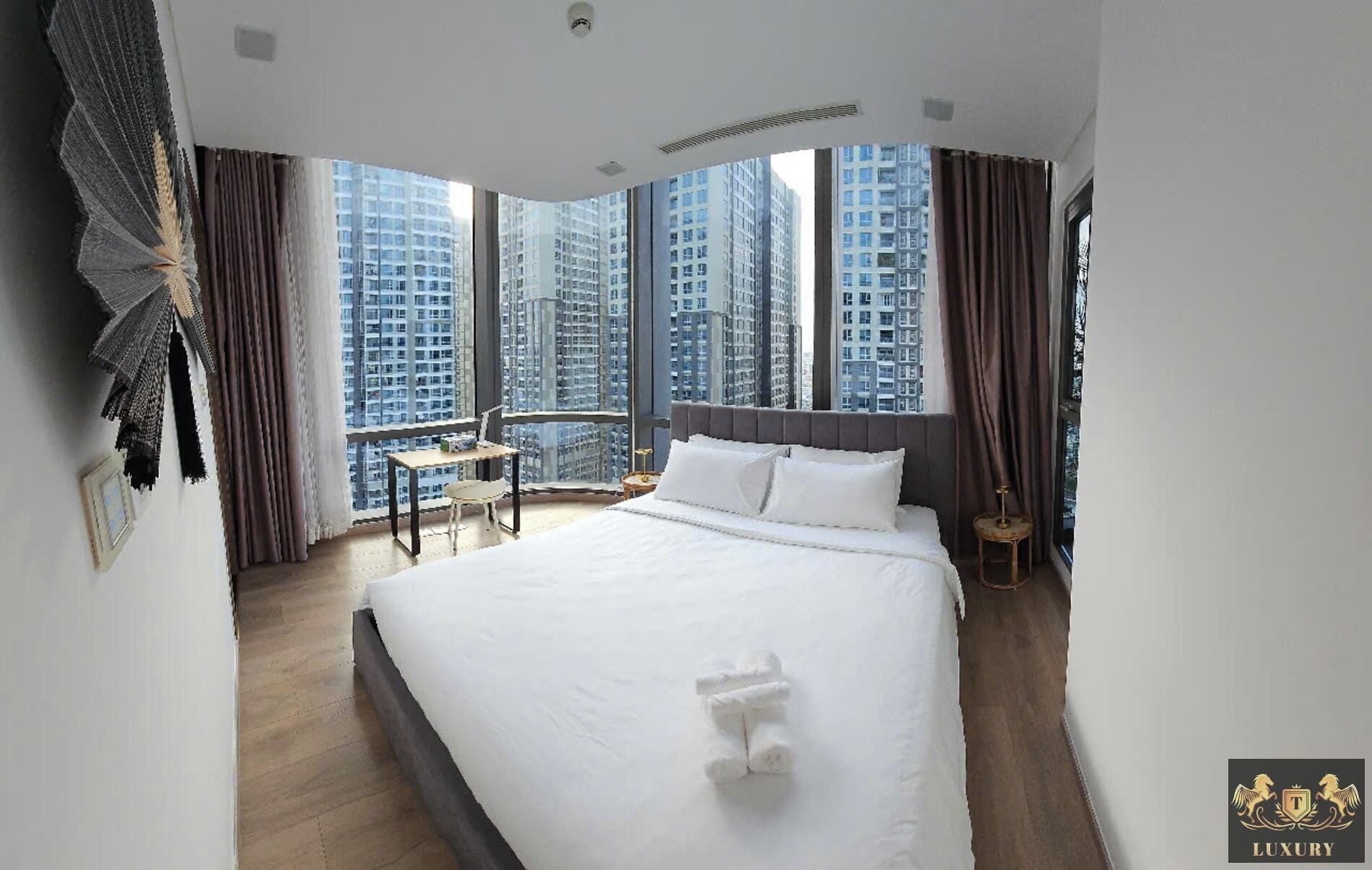 Habitación de hotel con una cama grande, un escritorio pequeño, una silla y vistas de edificios altos a través de amplios ventanales.