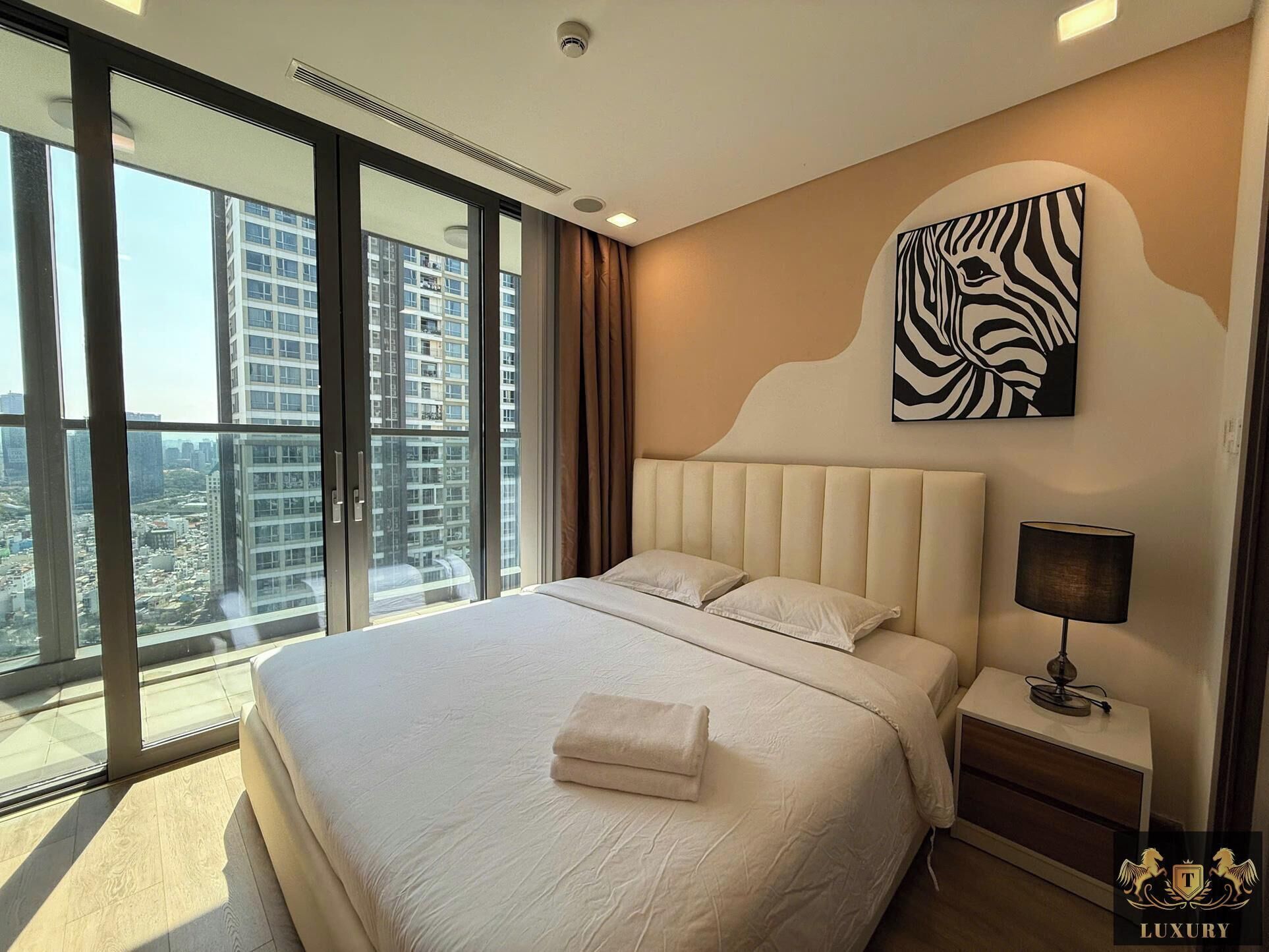 A modern hotel room with a large bed, a nightstand, a lamp, and a city view through floor-to-ceiling windows.