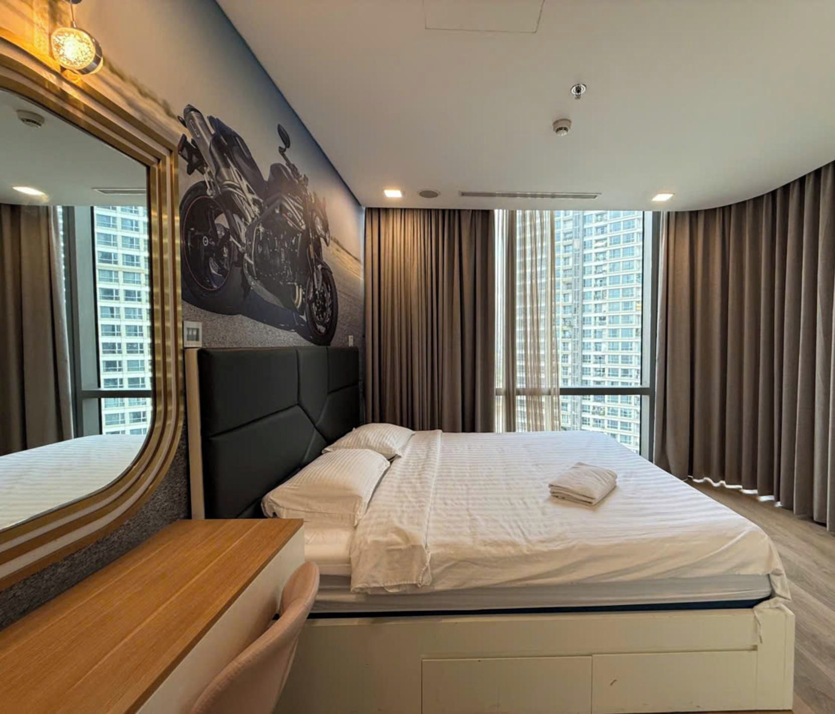 A hotel room with a large bed, a headboard, a wooden desk, a mirror, and a view of tall buildings through the windows.